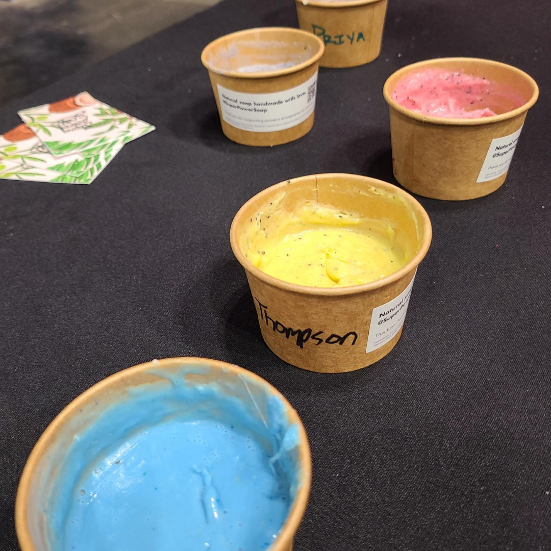 Super Power Soap - celebrate, relax, and rejuvenate with a team building workshop for your church group. Image of colorful workshop soaps in kraft paper cups on a black table. Scented with essential oils. Shea butter Soap. Happy participants at a table at the George R. Brown Convention Center during the Ultimate Women's Expo.