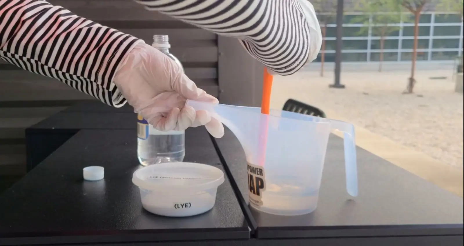 Evidence Image: Safety is taught as part of the process, including clothing and workspace choices. Gloved soapmaker preparing a labeled lye solution outdoors with safety equipment